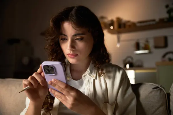 A young woman relaxes at home, intently focused on her smartphone this evening. — Stock Photo