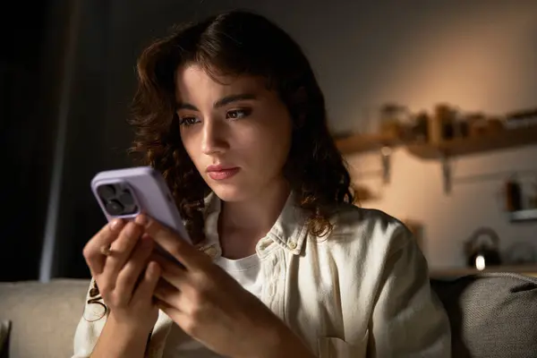 A beautiful young woman sits comfortably at home, deeply engaged with her phone, lost in thought. — Stock Photo