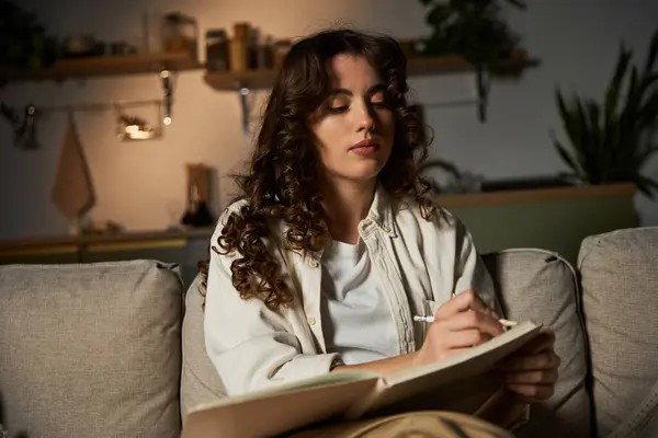 A young woman sits on a sofa, creating art under the soft glow of evening lights. — Stock Photo