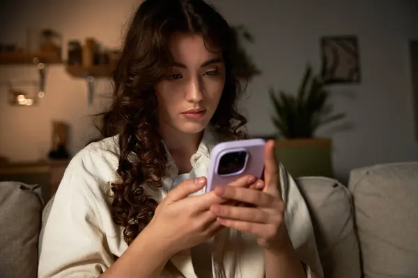 A young woman relaxes at home, focusing on her smartphone during a peaceful evening. — Stock Photo