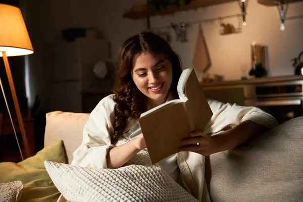 Eine junge Frau lächelt, während sie auf einer gemütlichen Couch liest, die zu Hause in warmes Licht getaucht ist. — Stock Photo