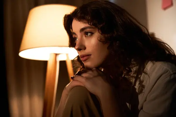 A beautiful young woman sits in a softly lit room, thoughtfully gazing away in quiet contemplation. — Stock Photo