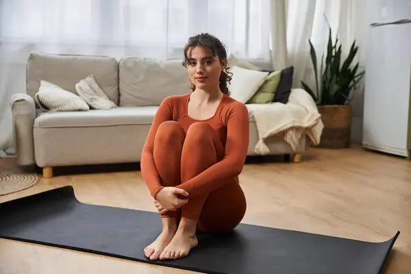 Una mujer joven se sienta en una postura serena en una esterilla de yoga en casa, encarnando la paz y el enfoque. — Stock Photo