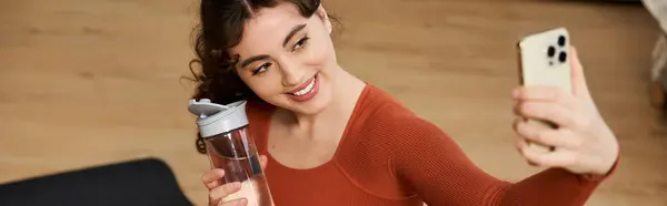 Young woman with a water bottle smiles while taking a selfie in her cozy home gym. — Stock Photo