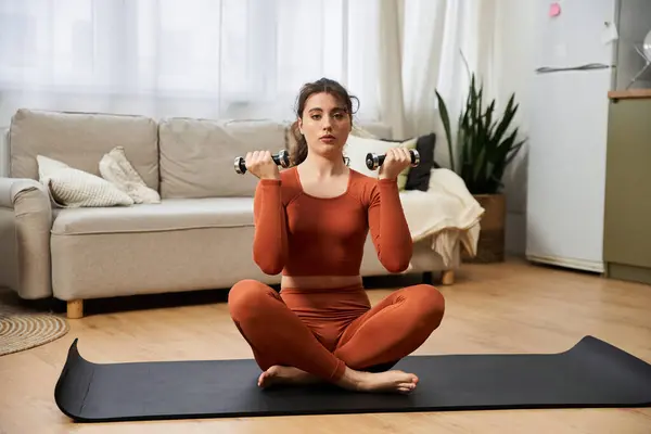 A beautiful young woman practices strength training with dumbbells while seated on a mat at home. — Stock Photo