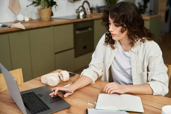 In a cozy kitchen, a beautiful young woman diligently works on her laptop, taking notes. — Stock Photo