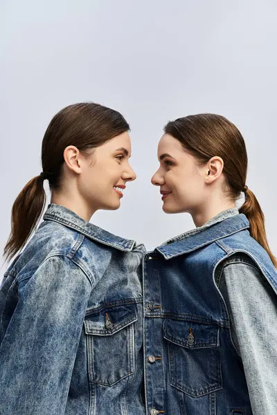 Two sisters smile at each other in matching denim jackets against a soft backdrop. — Stock Photo