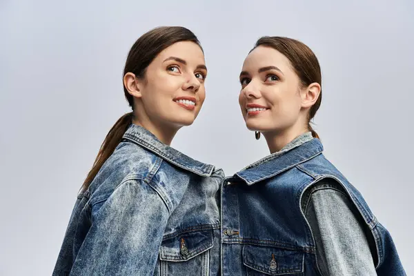 Identical sisters smile joyfully while wearing matching denim jackets against a light backdrop. — Stock Photo