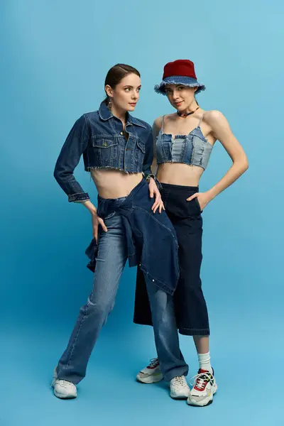 Two stylish sisters in trendy denim pose confidently against a bright blue backdrop. — Stock Photo