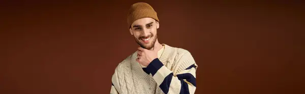 Un giovane uomo posa in un berretto alla moda ed elegante maglione su uno sfondo marrone caldo. — Foto stock