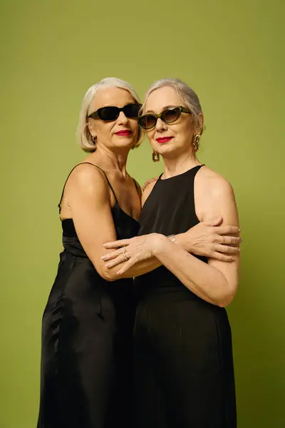 Two fashionable women embrace lovingly, radiating style and elegance against a green backdrop. — Stock Photo