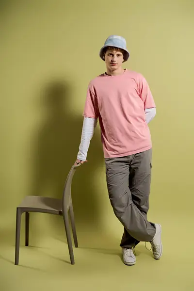 Dressed in a trendy pink tee and casual pants, a young man leans on a chair with style. — Stock Photo