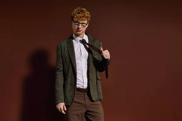 Un joven ajusta elegantemente su corbata mientras posa con confianza sobre un rico telón de fondo marrón. — Stock Photo