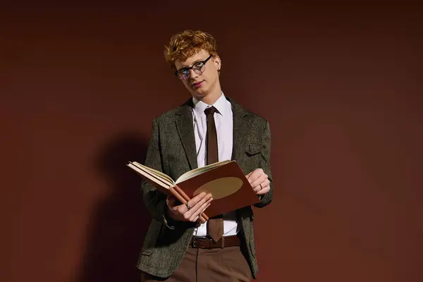 A young, fashionable man stands confidently, engrossed in a book, set against a rich brown backdrop. — Stock Photo