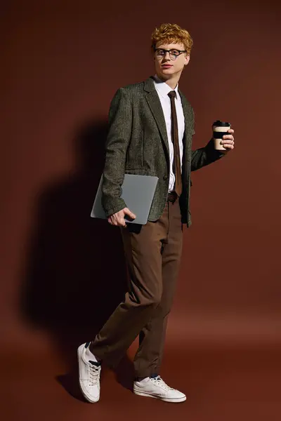 A young, fashionable man carries a laptop and coffee while exuding confidence in a modern office. — Stock Photo