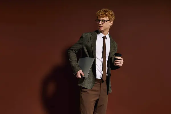 A fashionable young man dressed in a stylish blazer carries a laptop and coffee, exuding confidence. — Stock Photo