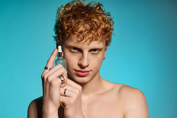 A fashionable young man holds a skincare serum, exuding confidence in a striking blue setting. — Stock Photo