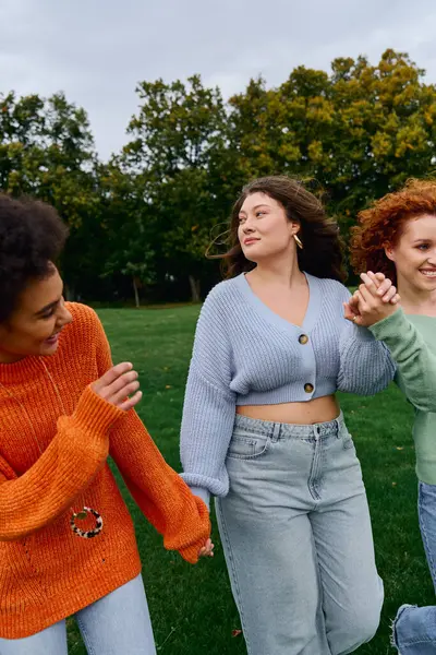 Three stylish friends stroll hand in hand through a lively spring park, filled with joy. — Stock Photo