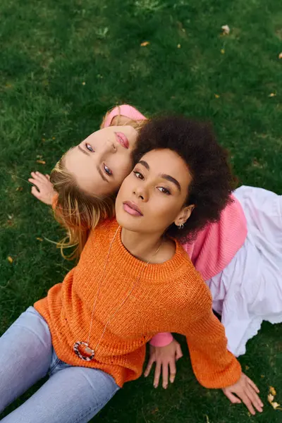 Two fashionable women relax on lush green grass, showcasing confidence and unique styles in spring. — Stock Photo