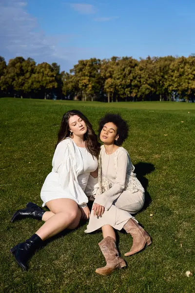 Two confident women relax on the vibrant green grass, showcasing their fashionable spring outfits. — Stock Photo
