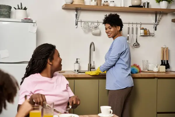 Dos alegres miembros de la familia comparten la risa durante el desayuno mientras un tercero se prepara en el fregadero. - foto de stock