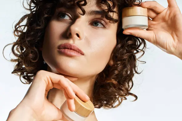 Young woman showcases elegant skincare jars with defined curls and a soft expression. — Stock Photo