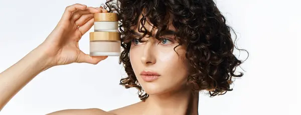 A beautiful young woman holds elegant skincare jars, radiating confidence in a calm space. — Stock Photo