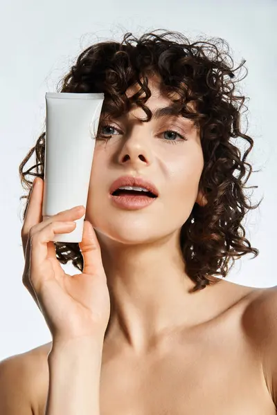 A young woman with curly hair holds a skincare product, radiating confidence in bright light. — Stock Photo