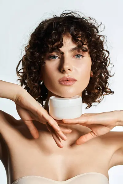 A beautiful young woman elegantly poses with a skincare jar beneath her chin, exuding charm. — Stock Photo