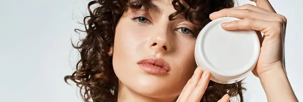 Una giovane donna con i capelli ricci mette in evidenza il suo prodotto di cura della pelle in luce morbida. — Foto stock