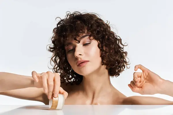 A young woman with curly hair gracefully opens a cosmetic jar, focusing on her beauty routine. — Stock Photo