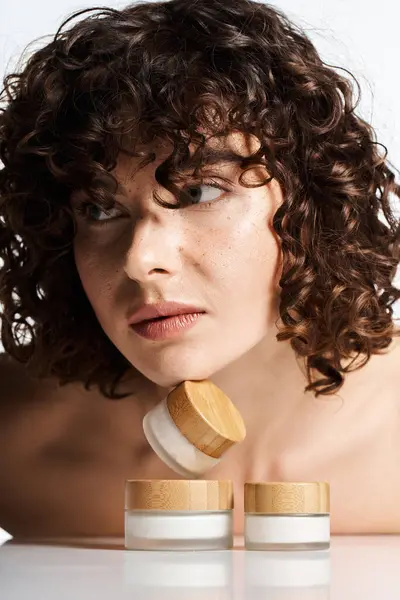 A young woman with curly hair highlights her beauty beside elegant skincare jars. — Stock Photo