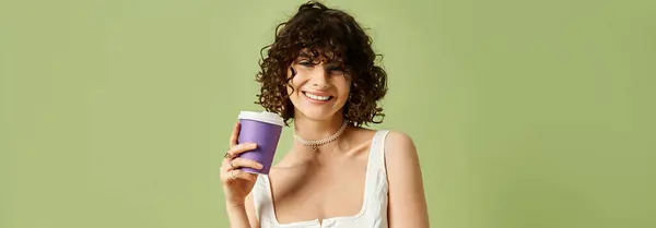 A young, beautiful woman with curly hair holds a coffee cup, radiating joy in a vibrant space. — Stock Photo