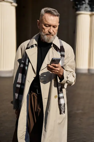 Un uomo anziano in un abbigliamento trincea alla moda cammina attraverso un ambiente urbano, incentrato sul suo smartphone. — Foto stock