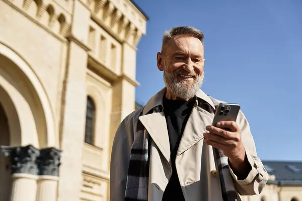 A fashionable senior man strolls through a vibrant city, checking smartphone with a beaming smile. — Stock Photo