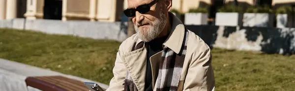 A stylish senior man sits casually on a bench in a vibrant cityscape, dressed in trendy attire. — Stock Photo