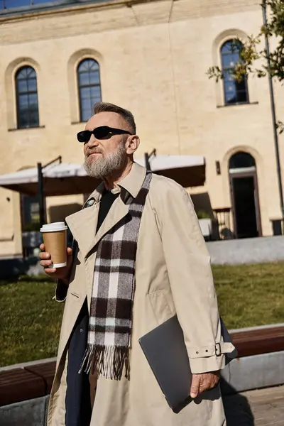 An attractive senior man wearing a trendy coat and sunglasses, holding a coffee cup and a laptop. — Stock Photo