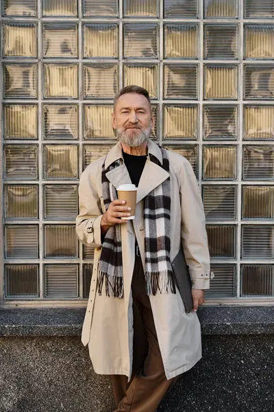 Un bell'uomo anziano in un trench alla moda e sciarpa si alza con fiducia, sorseggiando caffè. — Foto stock