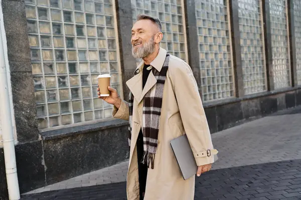 A fashionable senior man walks through urban setting, holding a coffee cup and a laptop. — Stock Photo