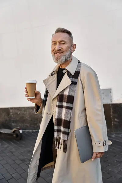 A dapper senior man strolls through the city, holding a coffee cup and a sleek laptop. — Stock Photo