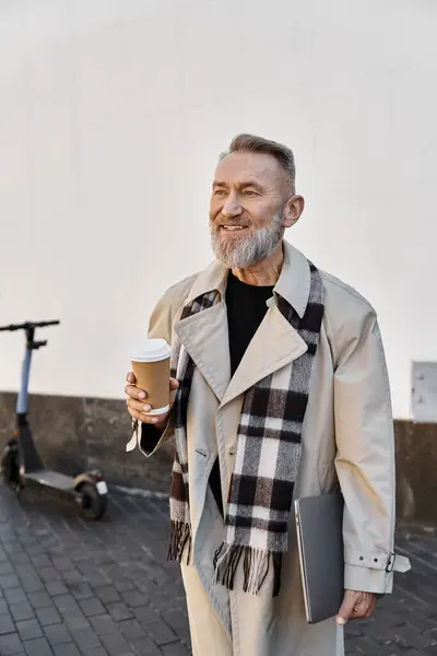 In una vivace zona della città, un uomo anziano alla moda si erge, con una tazza di caffè e un computer portatile. — Foto stock