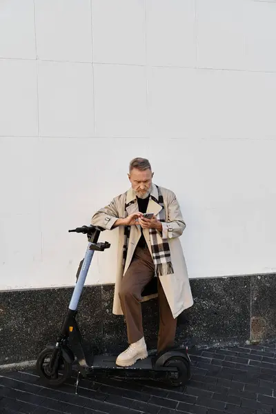 Un uomo anziano con un trench chic siede su uno scooter elettrico, assorbito nel suo telefono. — Foto stock