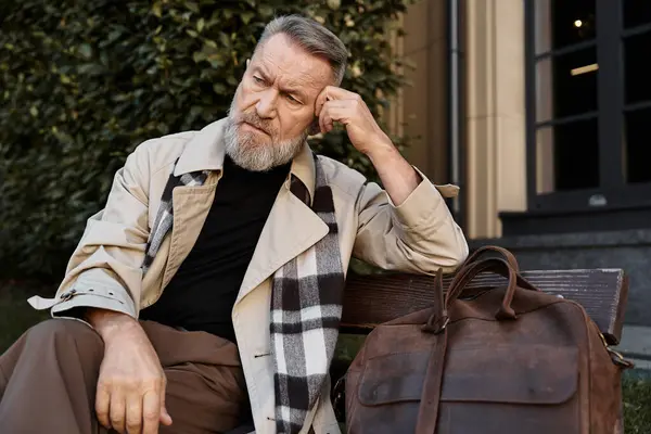Ein älterer Mann mit gepflegtem Bart sitzt nachdenklich auf einer Bank, umgeben von urbanem Grün.. — Stockfoto
