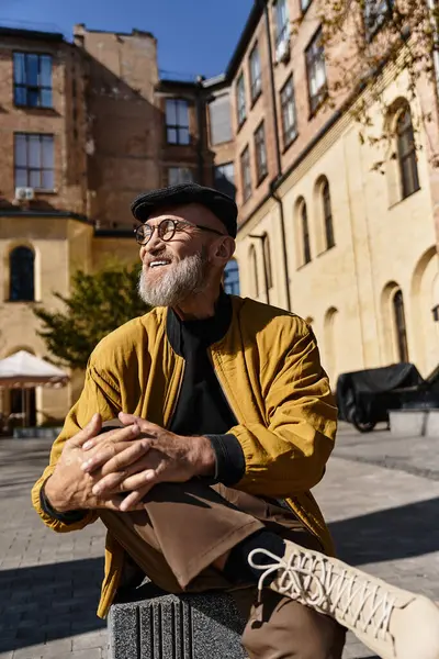 A handsome senior man sits comfortably in a chic city square, dressed in a trendy jacket and pants. — Stock Photo