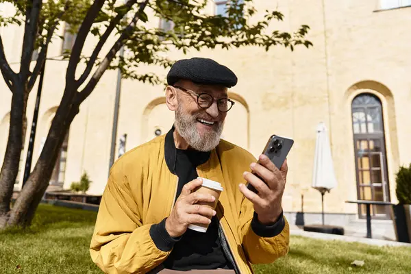 A fashionable senior man with a beard and glasses smiles while checking his smartphone. — Stock Photo