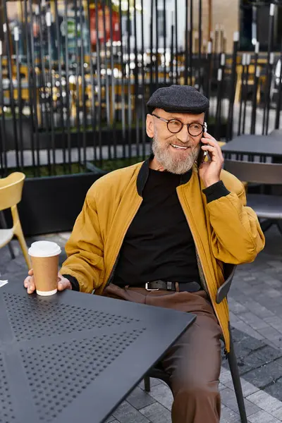 Um homem sênior senta-se na mesa de café ao ar livre, absorvido em uma conversa telefônica, segurando uma xícara de café. — Fotografia de Stock