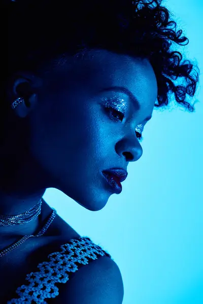 A stylish African American woman flaunts chic makeup under soft blue lighting. — Stock Photo