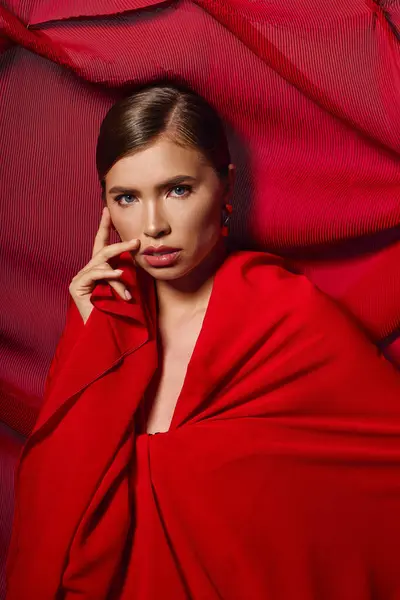 A stylish young woman poses gracefully with vibrant red fabric artfully arranged behind her. — Stock Photo