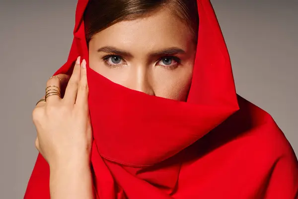 A stylish woman drapes a red shawl over her face, revealing her striking eyes in a chic setting. — Stock Photo