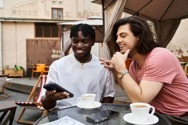 A handsome gay couple shares smiles and coffee at a cafe while using their phones. — Stock Photo